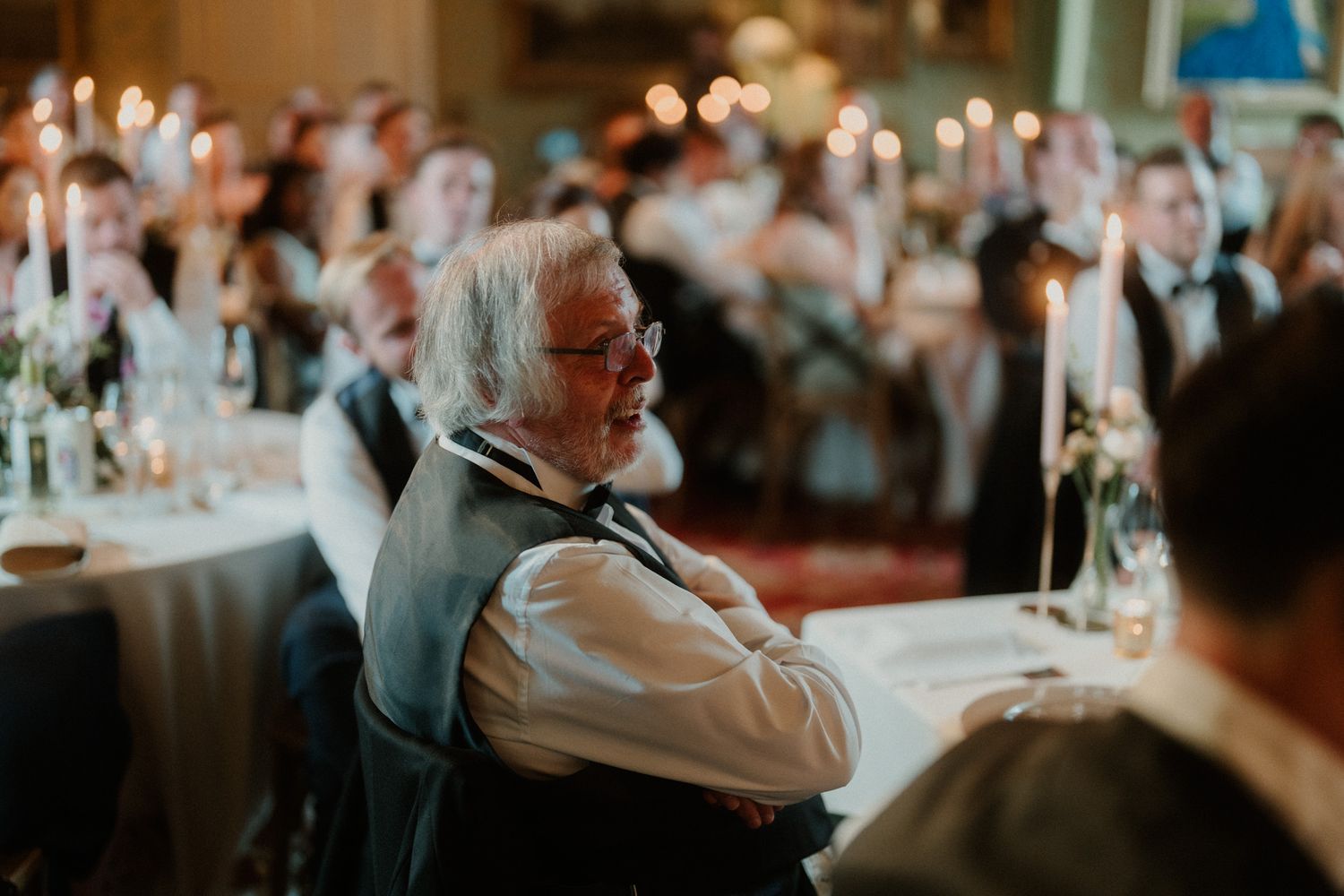 Smiling gray-haired man with glasses in a vest sits at a candlelit banquet table while guests dine in the warm, festive background.