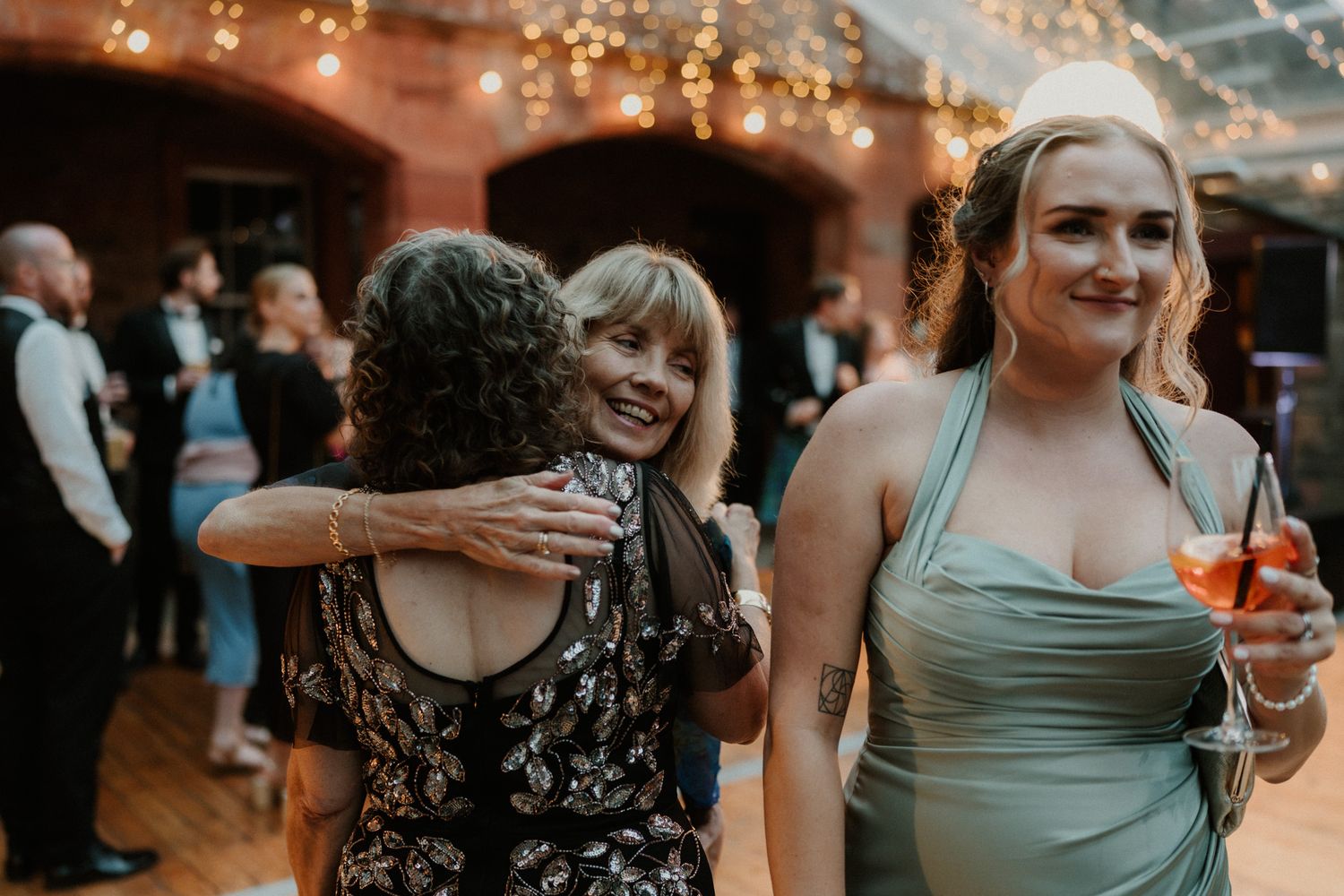 Two women embrace at an indoor wedding reception while a woman in a sage green dress holds a spritz cocktail and smiles nearby.