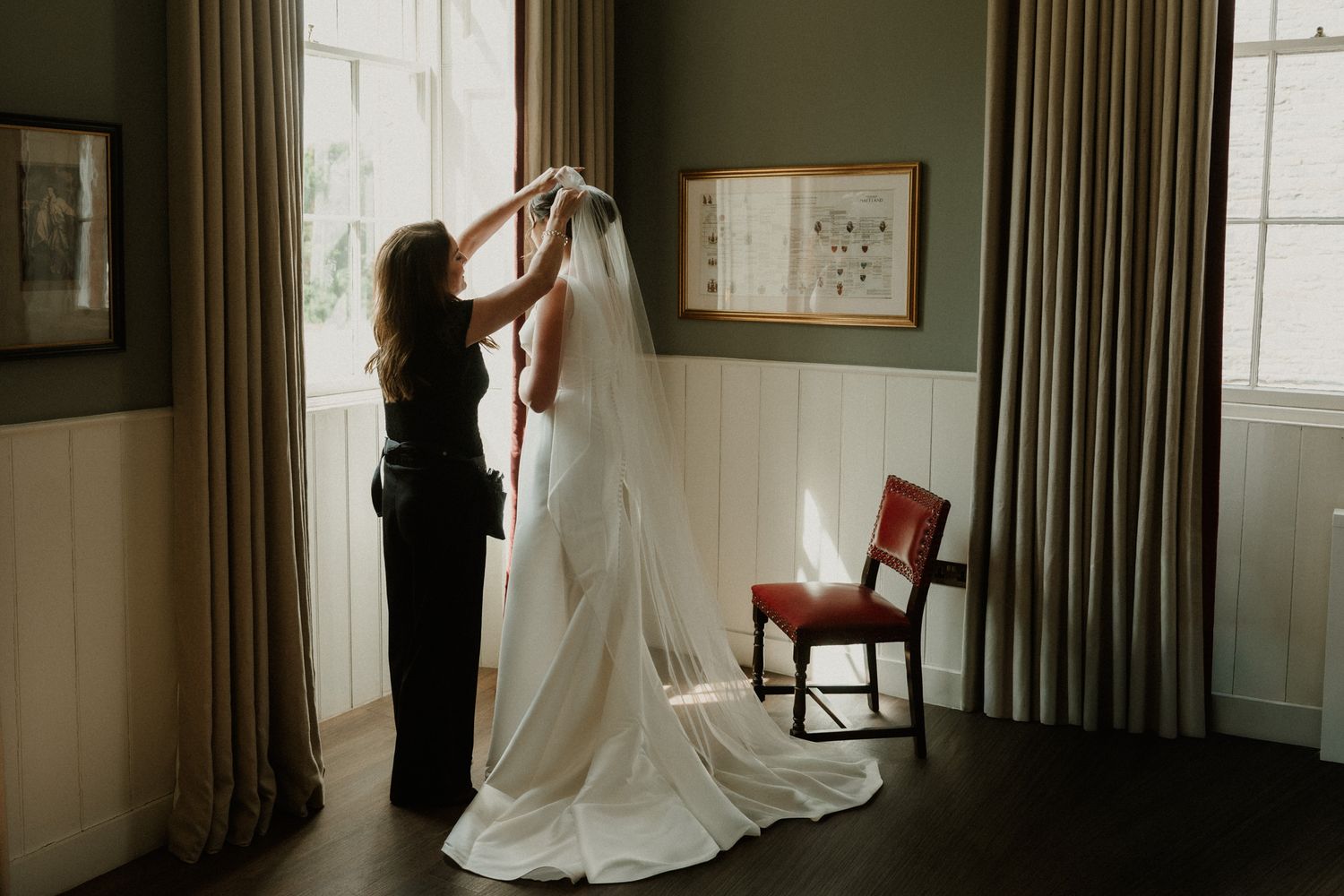 A woman in black adjusts the long veil of a bride in a white gown beside a sunlit window in an elegant room with a red chair.