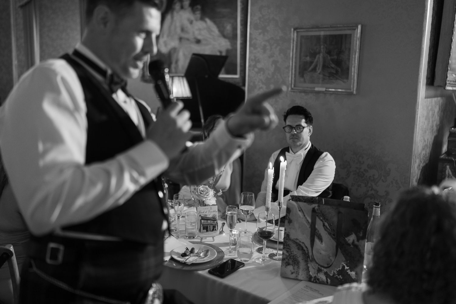 Man in a vest speaking into a microphone and pointing during a wedding reception toast, with guests seated at a table behind him.