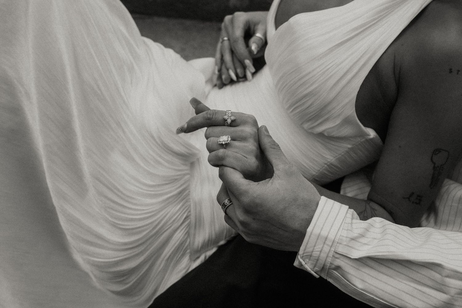 Black and white photo of hands holding, with a woman in a white dress and a man in a dress shirt, wearing rings.
