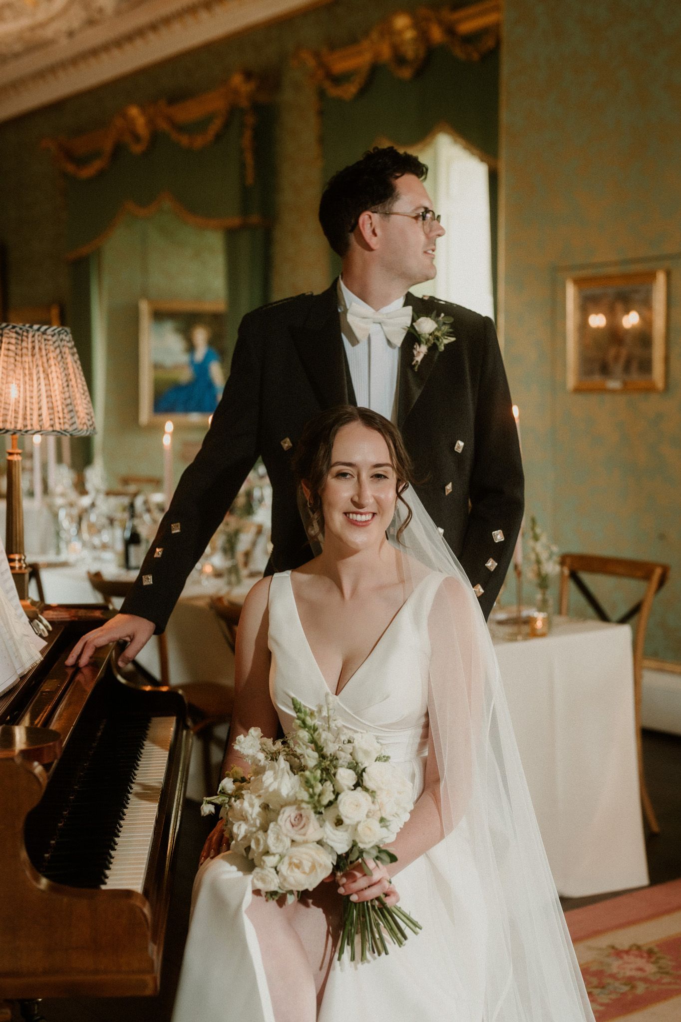 Smiling bride in a white gown holding a bouquet sits by a piano as the groom in a black jacket stands behind her in an elegant green room.