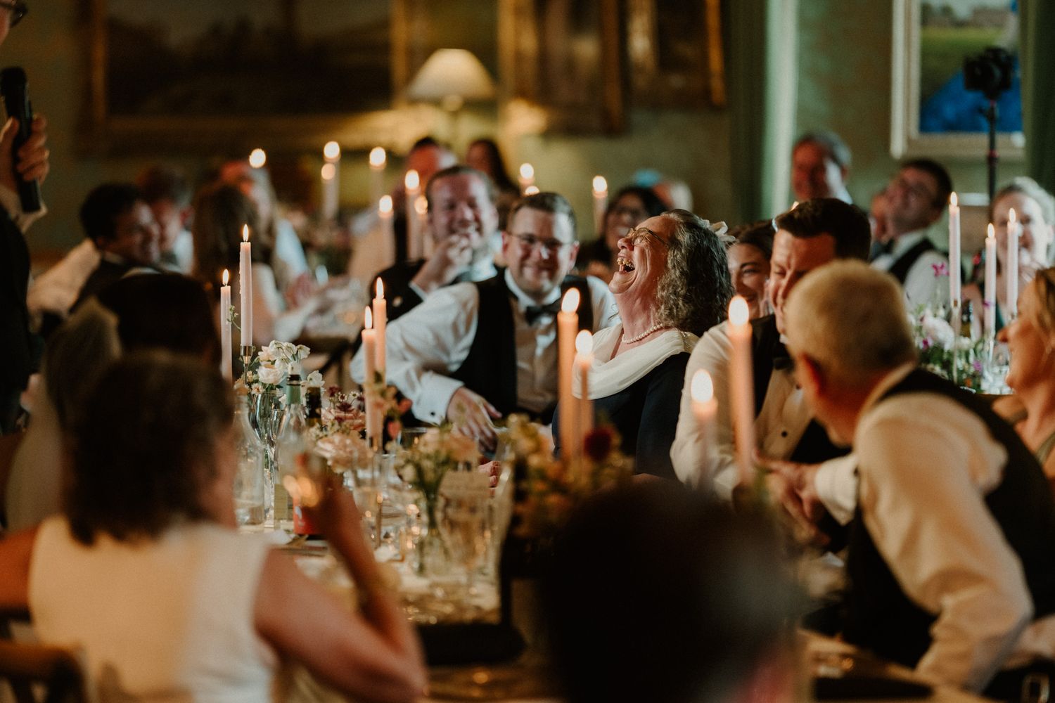 Wedding guests laugh and celebrate around a candlelit banquet table in an elegant, warmly lit reception room.