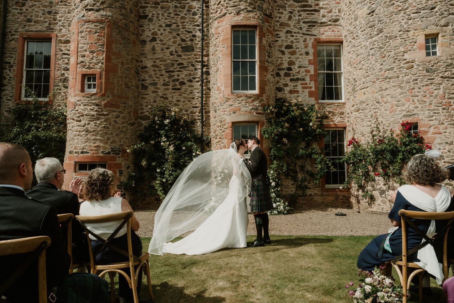 Bride in a white gown with a long flowing veil and groom exchange vows during an outdoor ceremony at a stone castle as guests watch.
