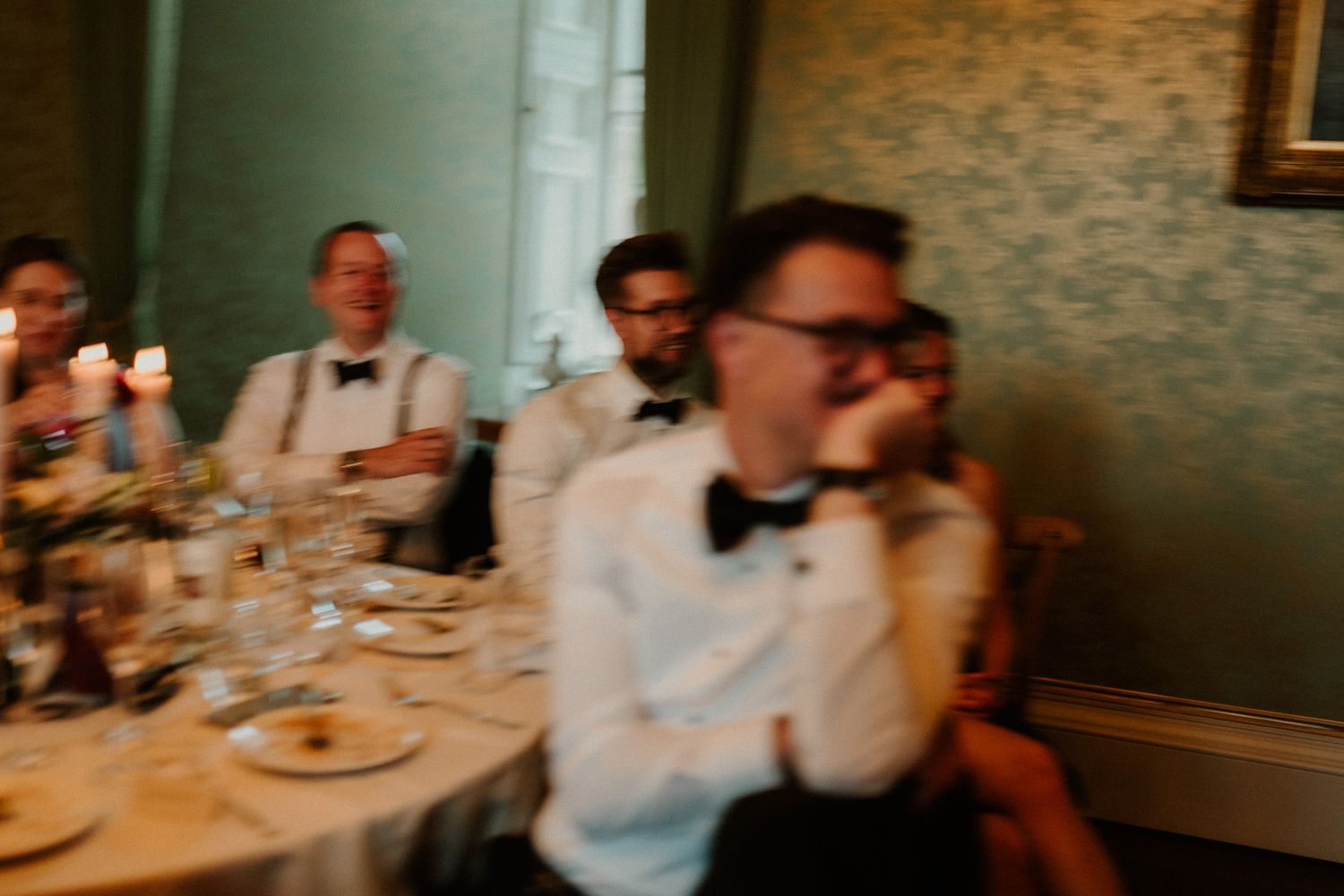 Blurry wedding reception scene with men in bow ties and white shirts seated at a candlelit table in a room with green wallpaper.