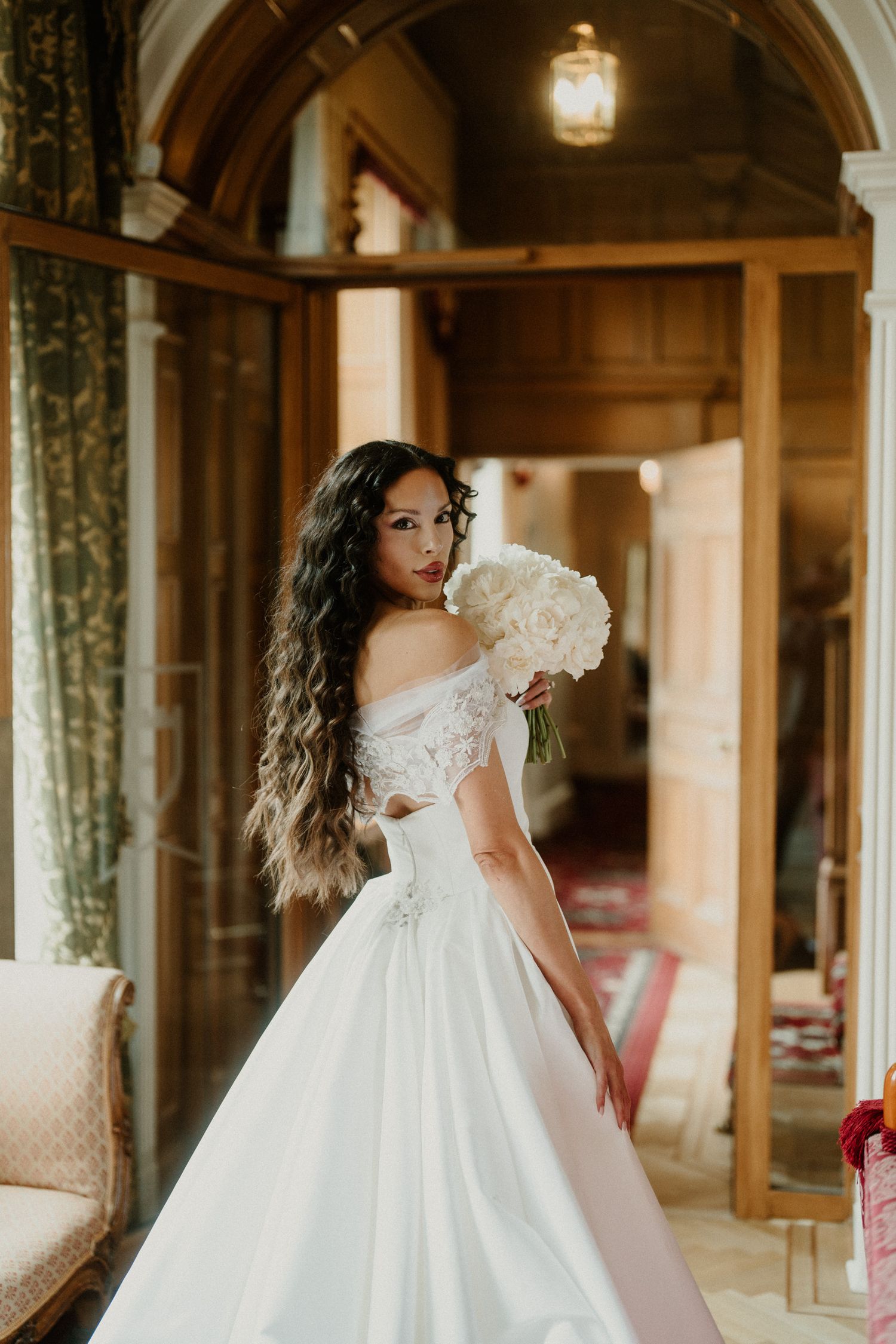 A bride in an off-shoulder wedding gown poses with white flowers in an elegant vintage-style interior.