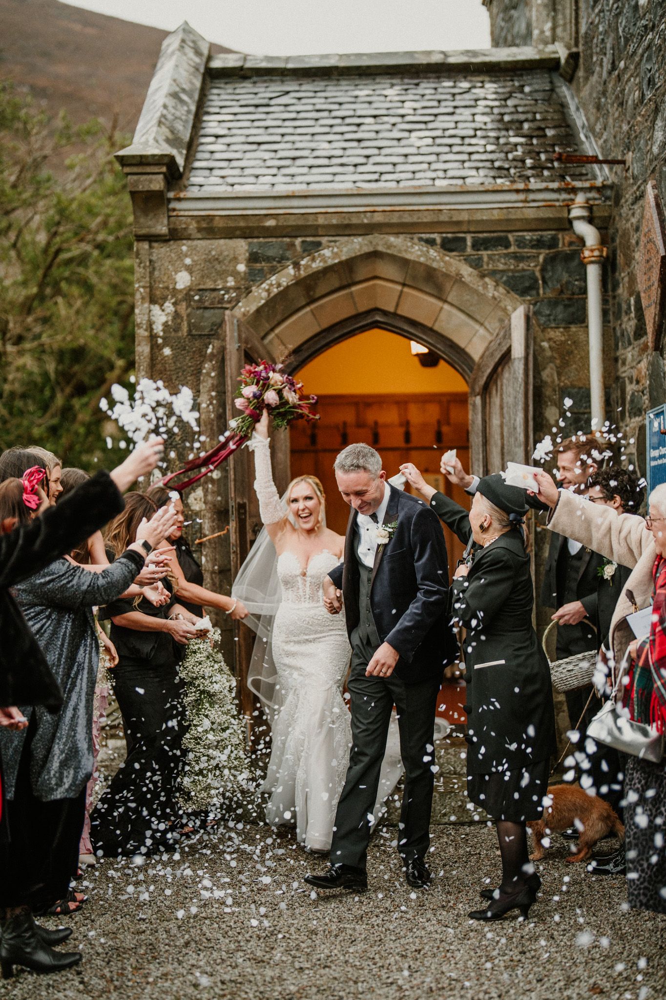 Wedding party exits church while guests throw white confetti in celebration.