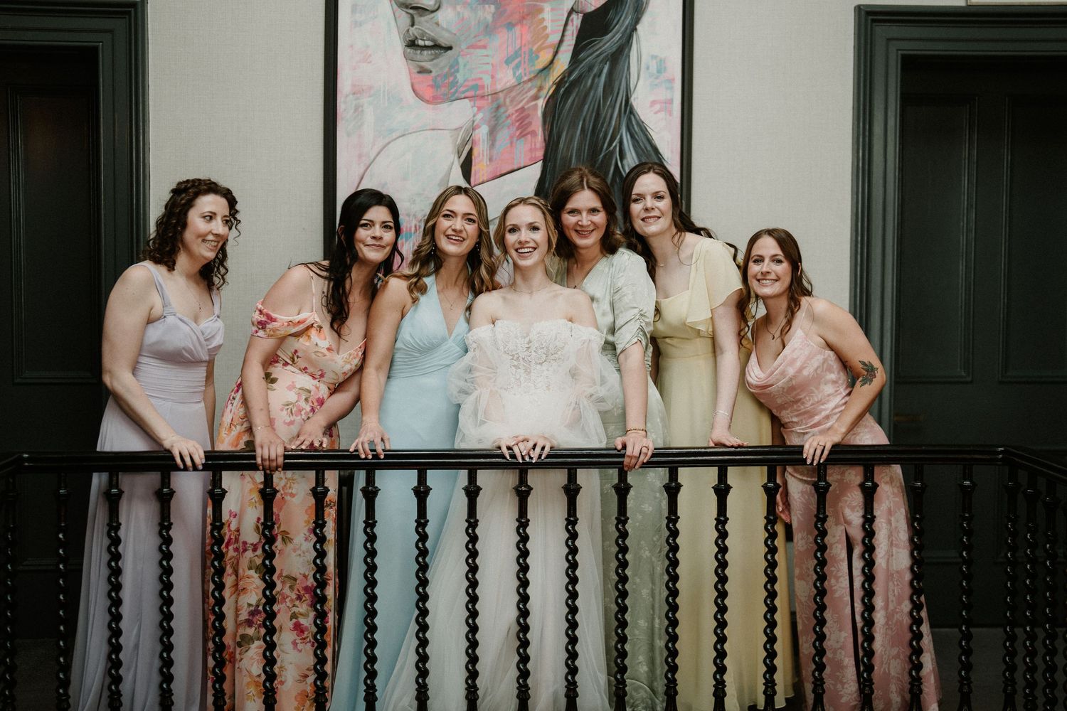 Wedding party in pastel dresses poses on balcony with vintage artwork backdrop.