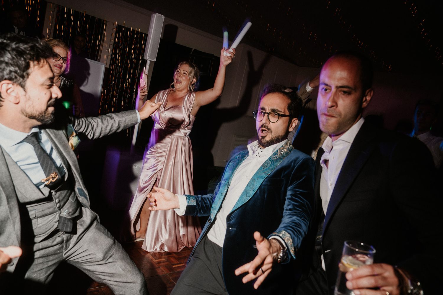 Wedding reception guests socializing and dancing in a dimly lit venue with decorative lighting.
