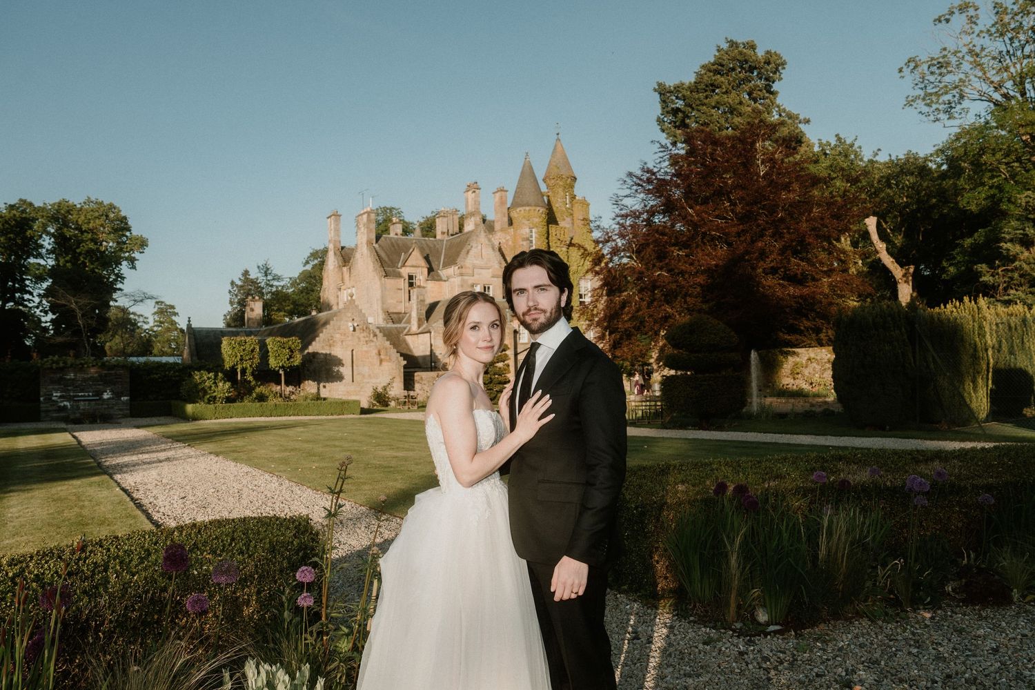 A romantic wedding couple poses in front of an ivy-covered stone castle during golden hour.