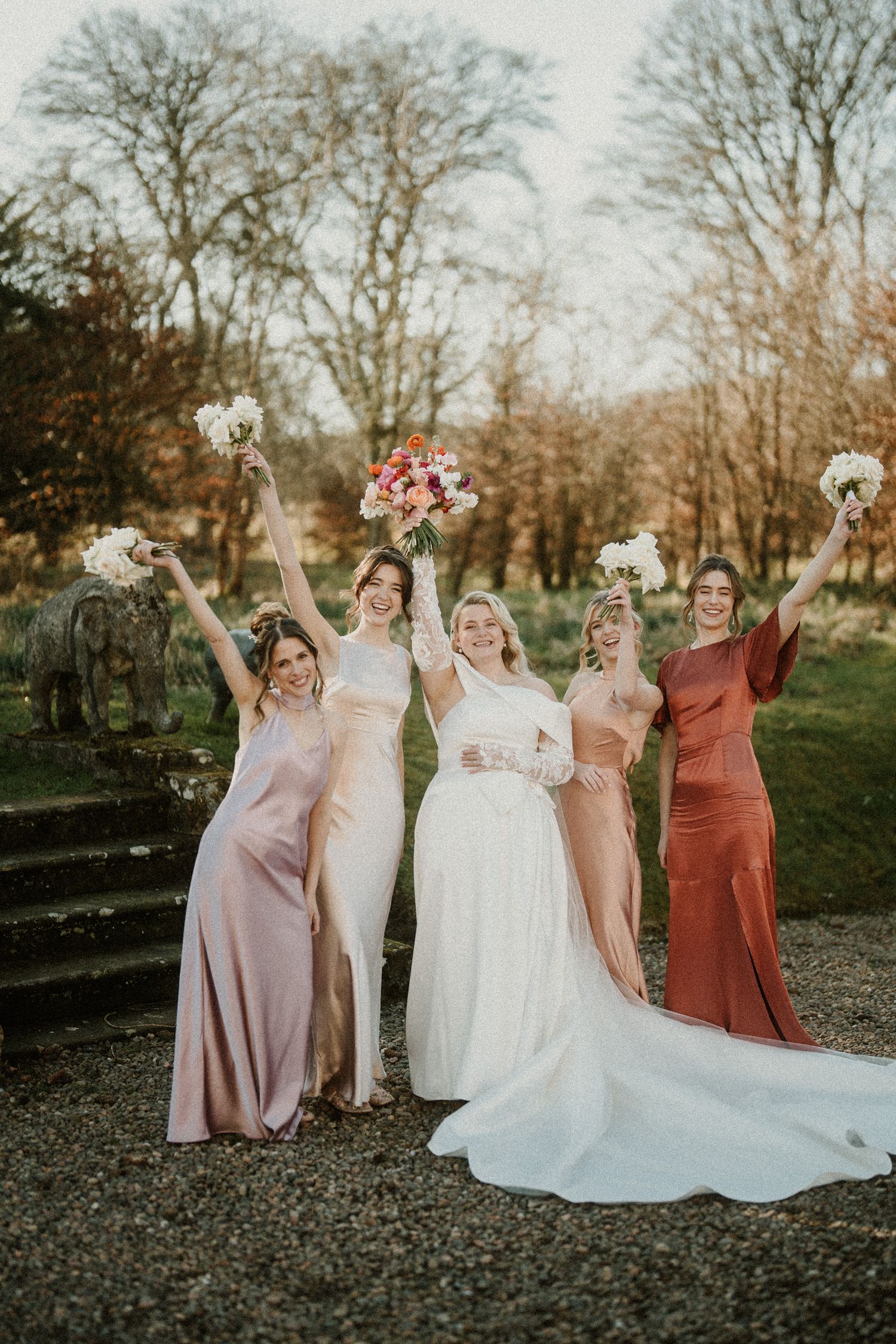 Bridal party in blush and rust colored dresses pose together outdoors during autumn wedding celebration.