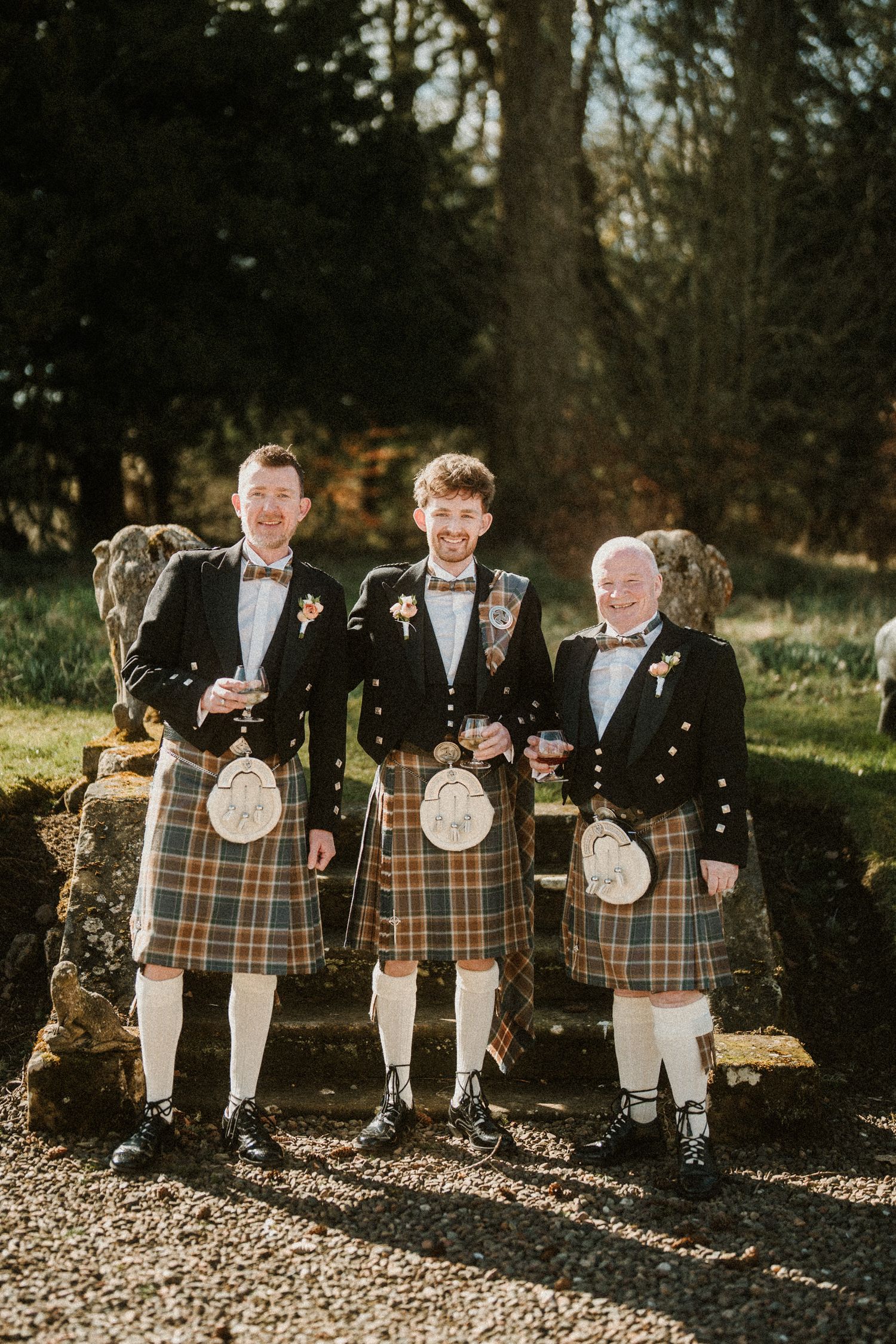 Three men in formal Scottish attire with kilts pose outdoors against a natural backdrop.