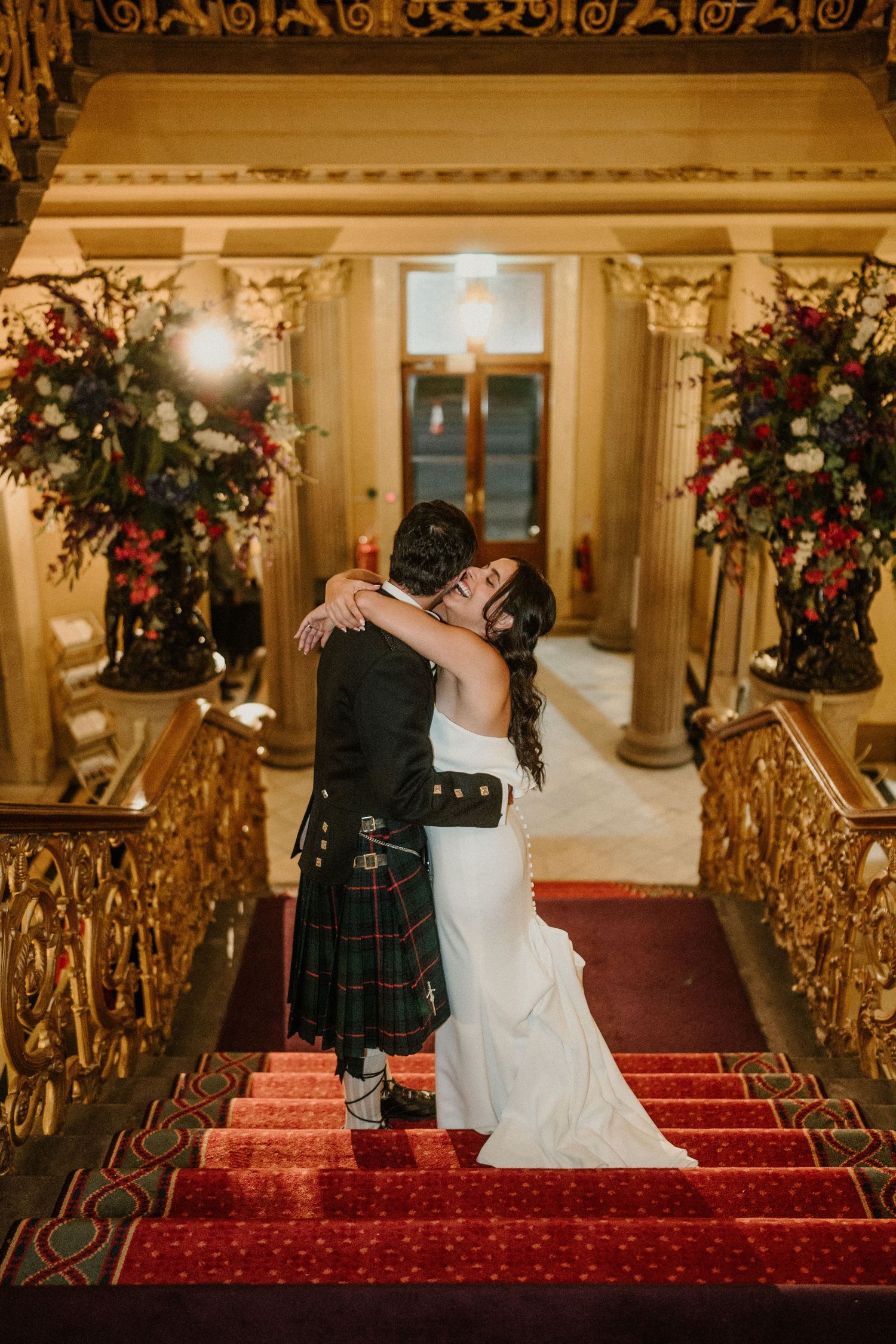 A couple shares a romantic moment on red-carpeted stairs in an ornate venue with gold railings.
