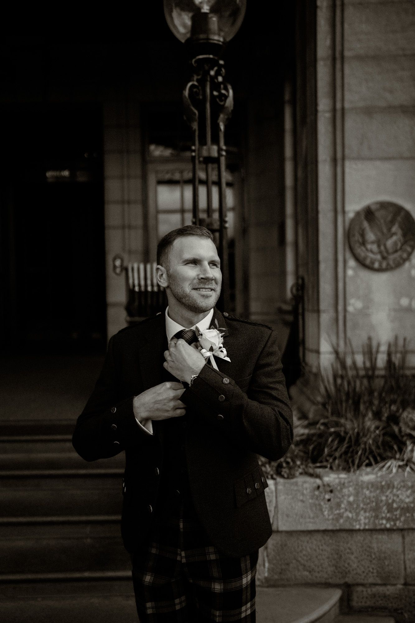 Groom in traditional Scottish kilt smiling outside stone building.
