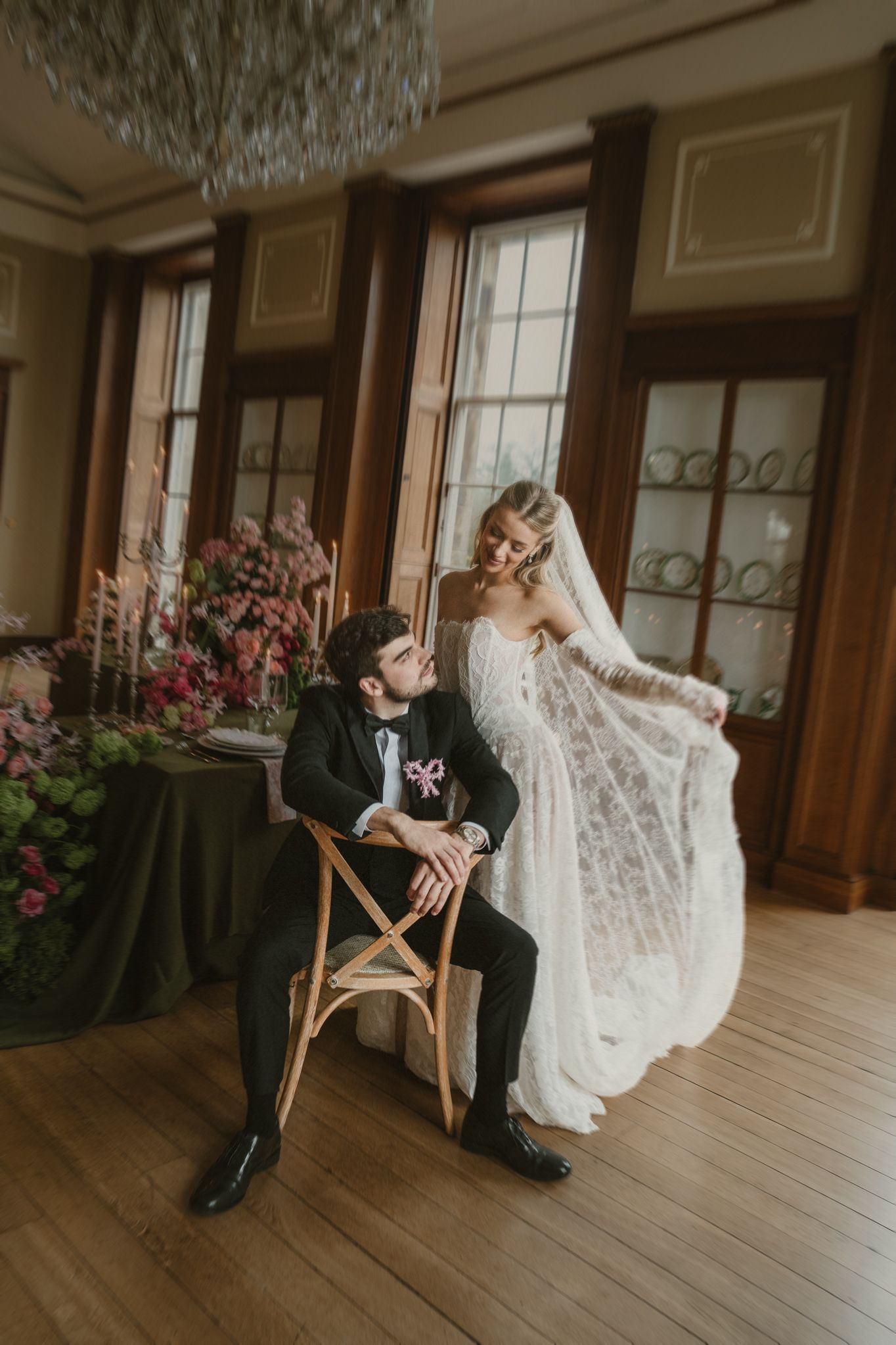 A couple in wedding attire shares a playful moment in a grand room with wooden floors and crystal chandeliers.