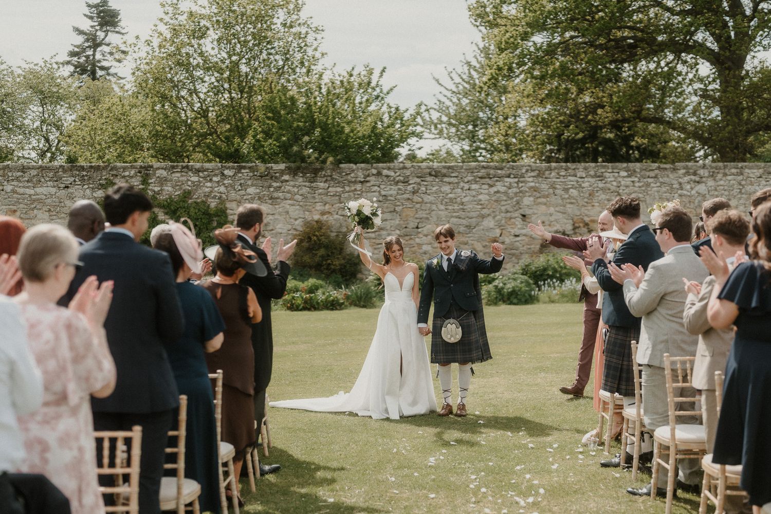 Bride and groom celebrate their outdoor Scottish wedding ceremony, groom in kilt, guests applauding along the aisle.