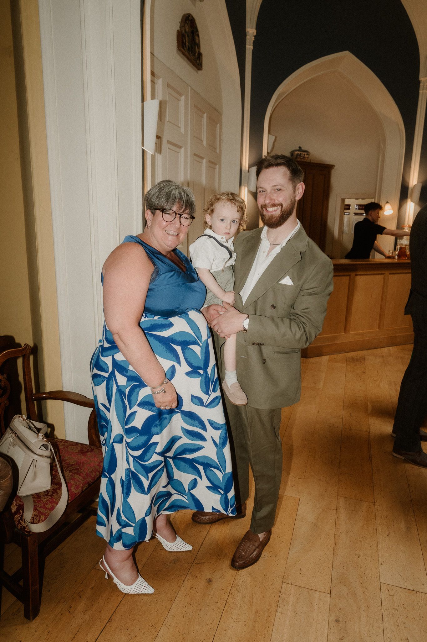 A woman in a blue floral dress and a man in a sage suit hold a toddler at an elegant indoor venue.