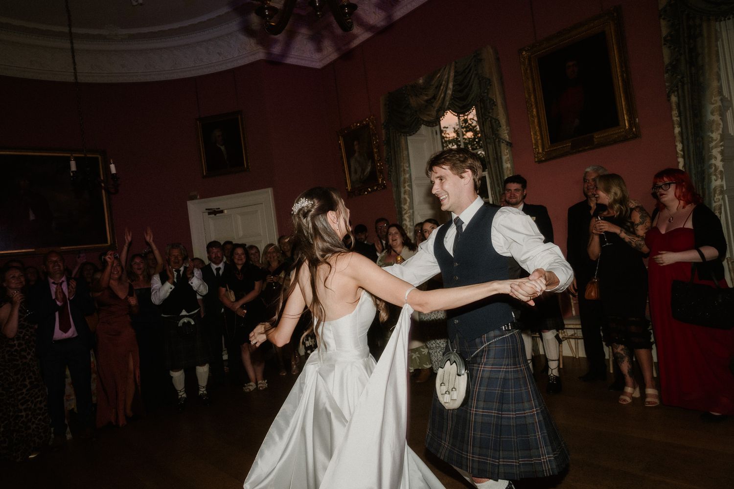 Bride and groom share first dance in Scottish castle ballroom, groom wearing traditional tartan kilt, guests celebrating in background.