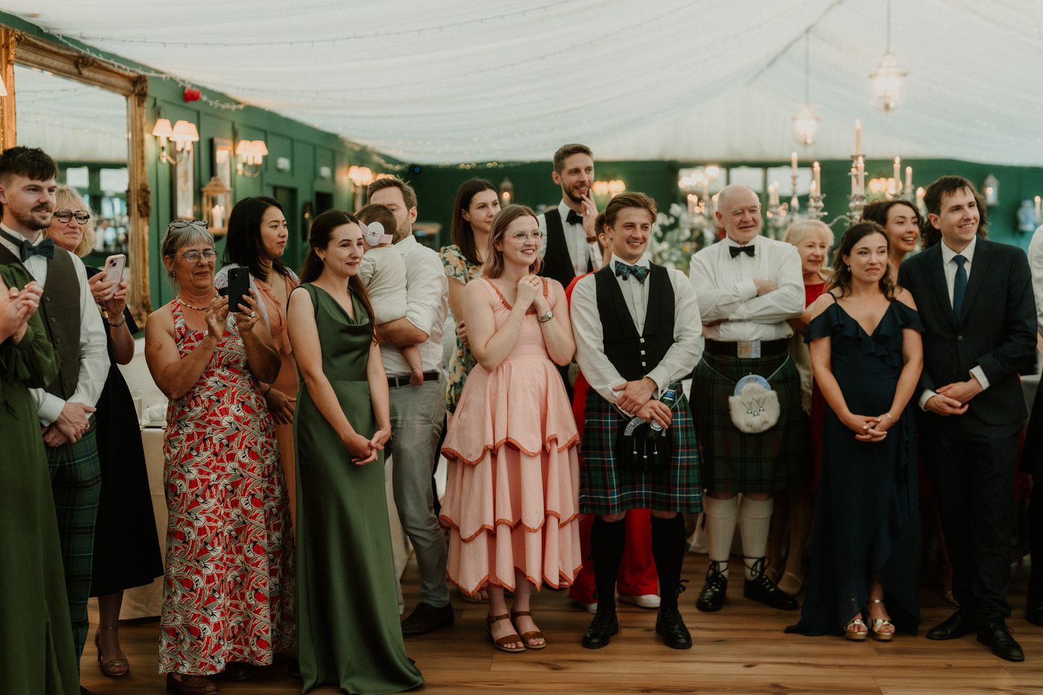 Wedding guests in formal attire, including kilts and evening gowns, applauding at an elegant marquee reception.