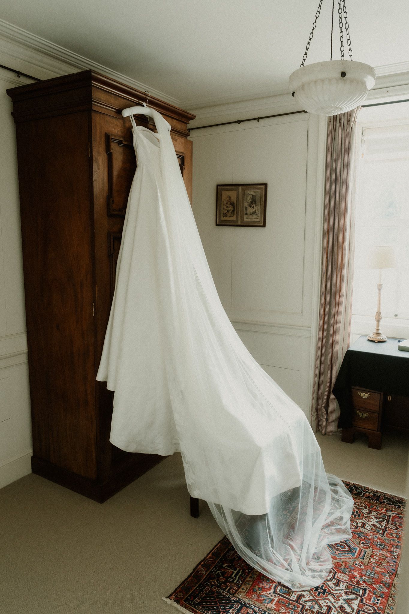 Elegant white wedding dress with long tulle train hanging on a wooden armoire in a classic bridal suite.