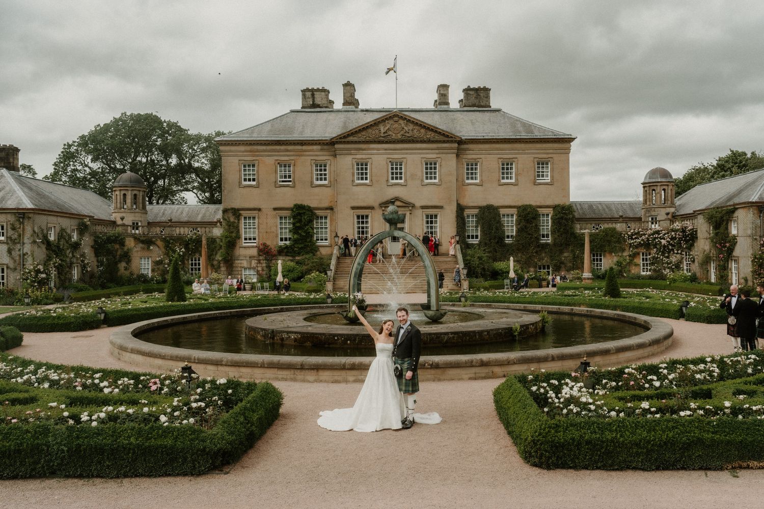 Bride and groom pose by a circular fountain in formal gardens of a grand Georgian country house estate.