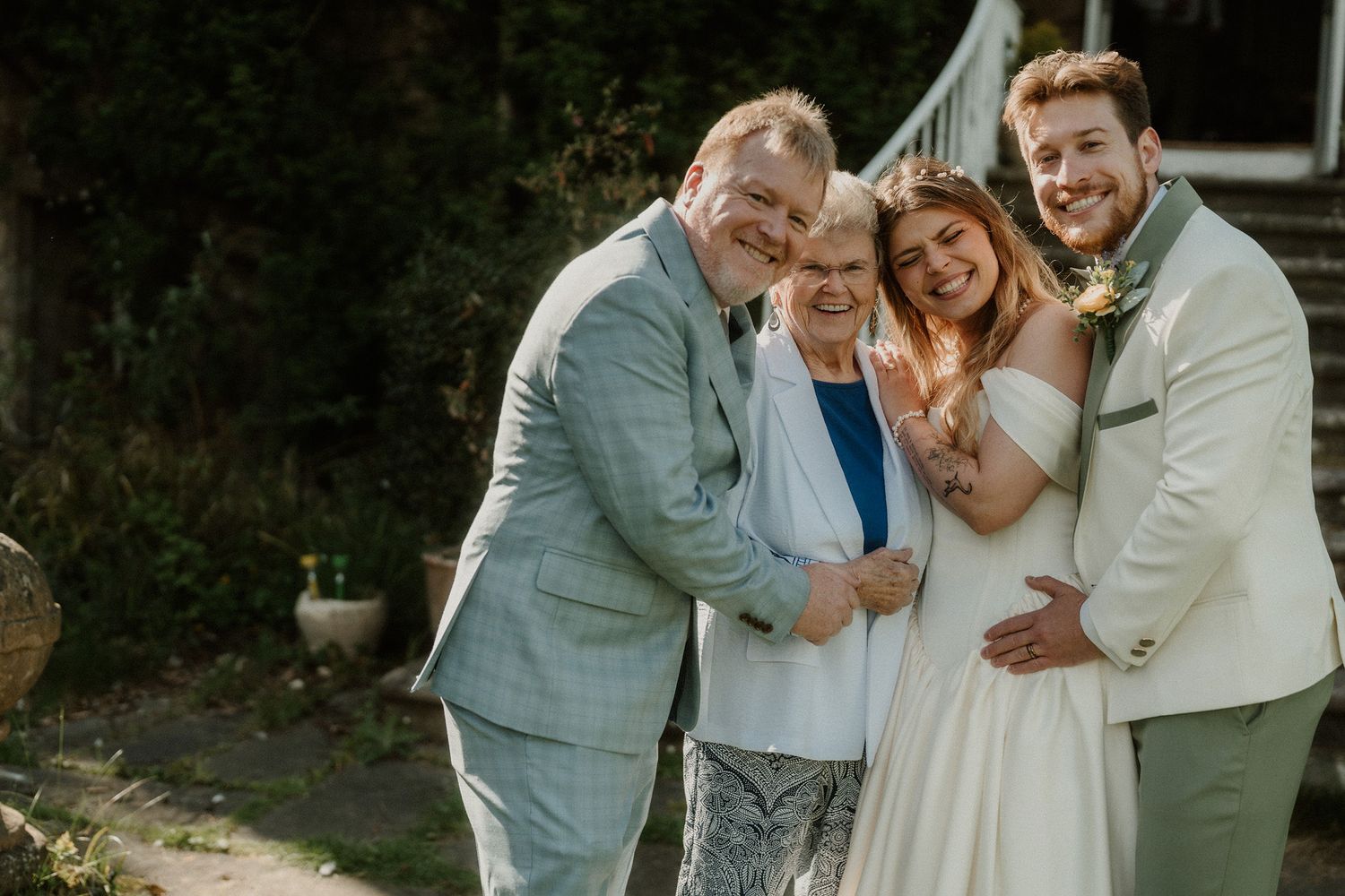 Wedding party of four smiling outdoors, bride in white gown with groom and family in sage green suits.