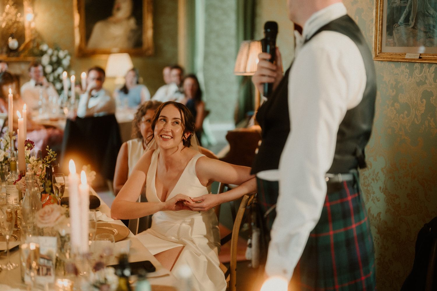 A smiling bride in a white gown sits at a candlelit table while a man in a kilt speaks into a microphone at a wedding reception.
