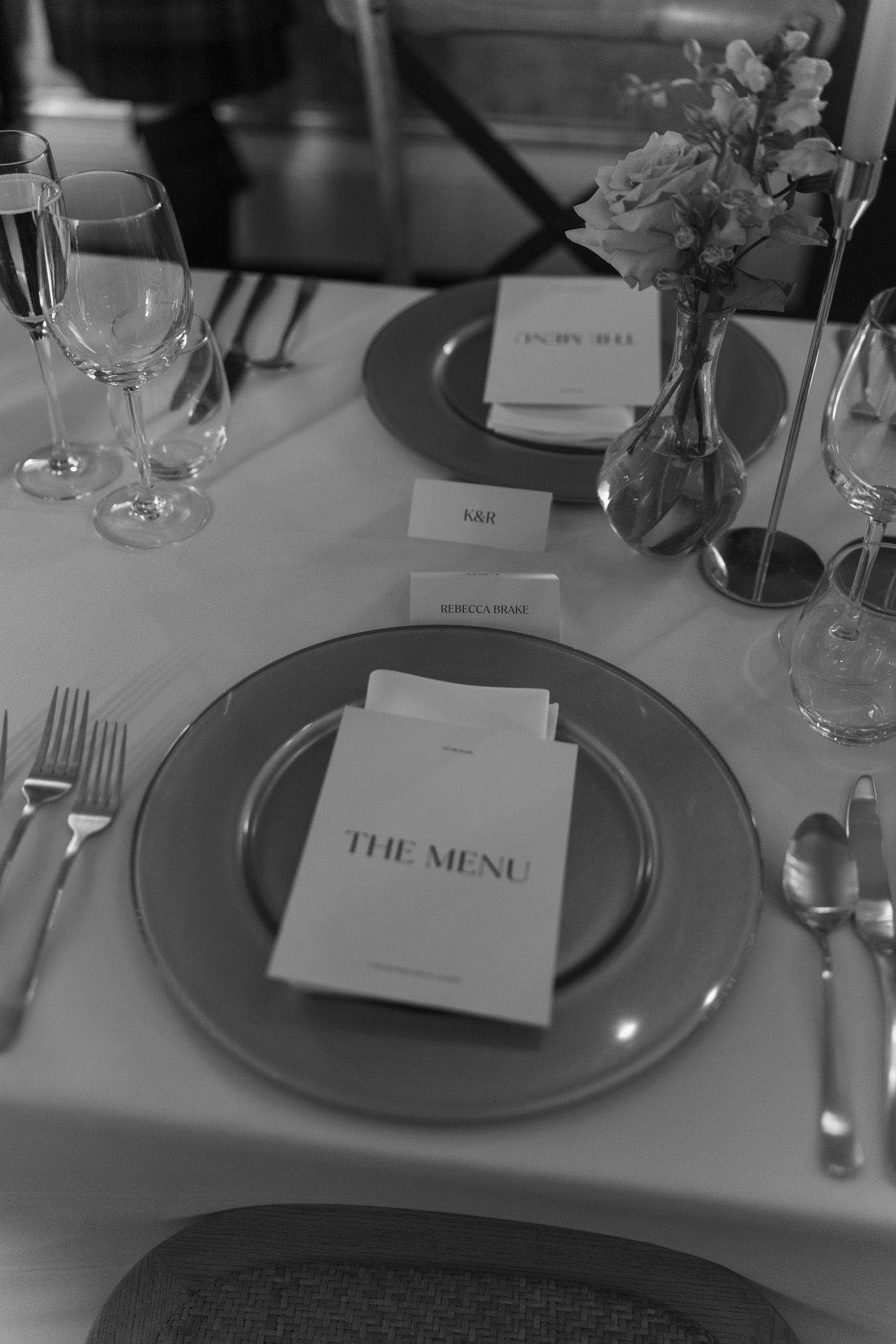Black and white photo of an elegant wedding table setting with a printed menu on a charger plate, glassware, flowers, and silverware.
