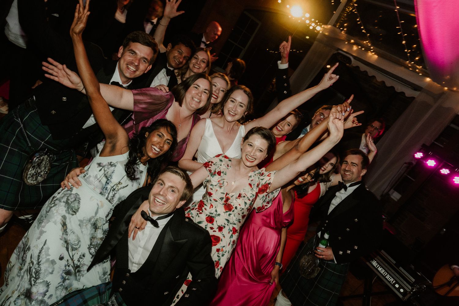 Joyful wedding guests in formal attire and floral dresses raise their arms and cheer together on a festive dance floor under party lights.