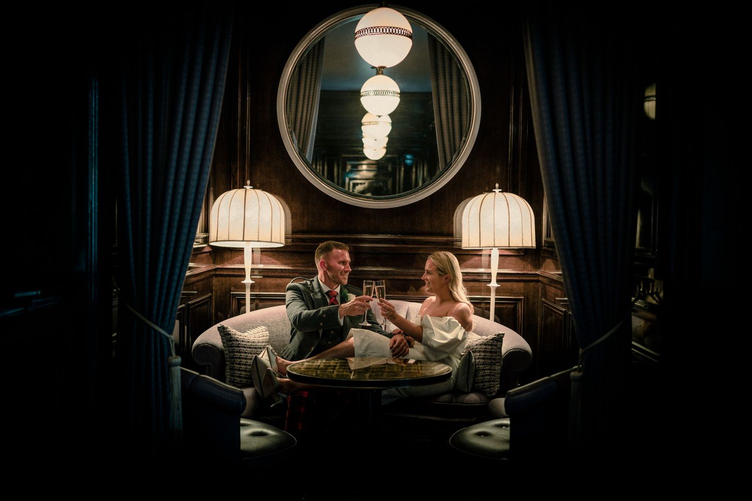 Two people relax on an elegant sofa beneath circular mirrors and pendant lights in a moody, luxurious setting.