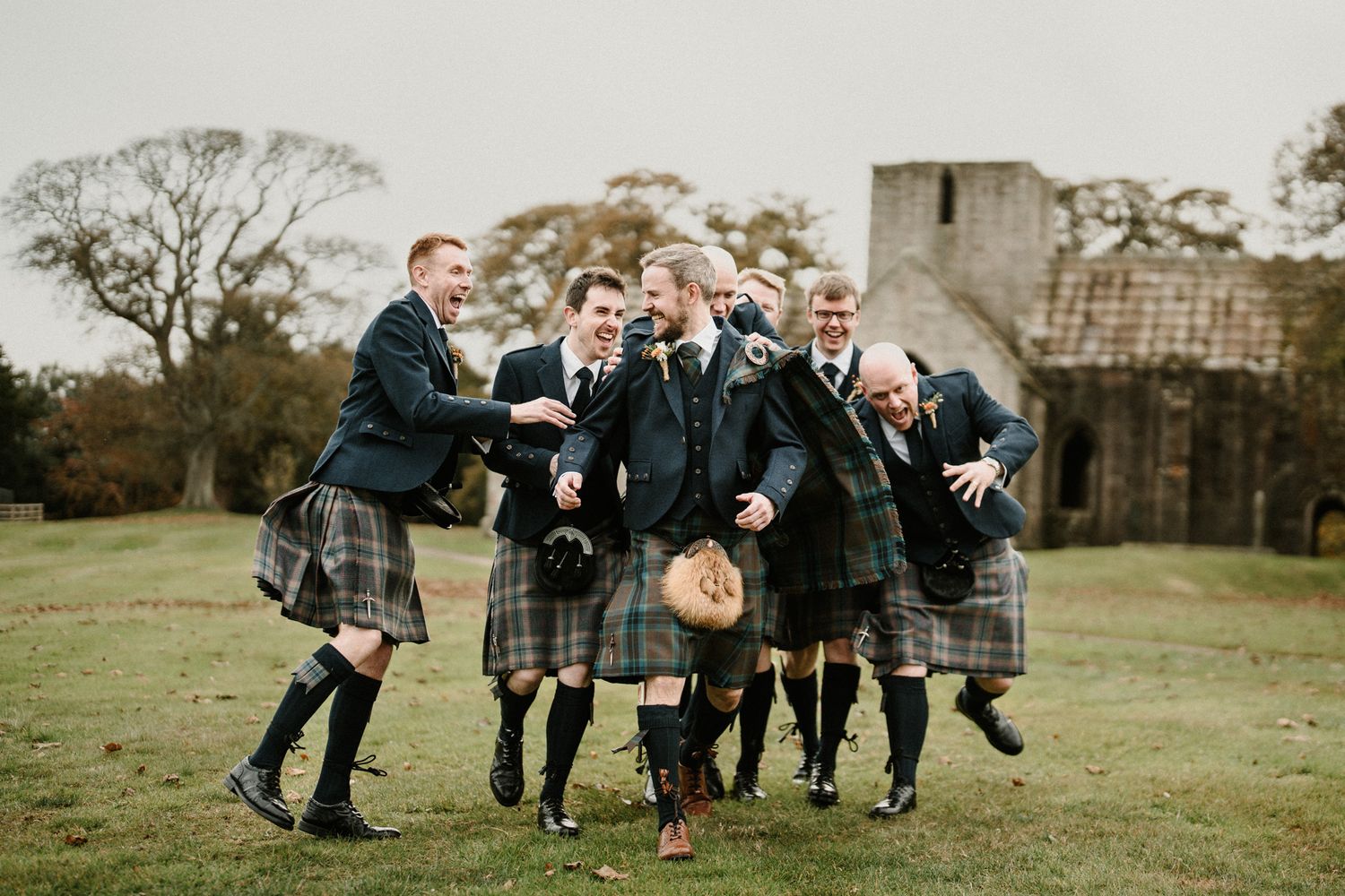 A group wearing traditional Scottish kilts walks across a grassy field near stone ruins.