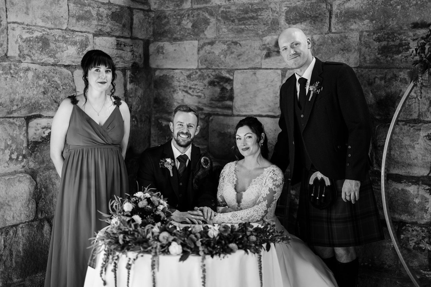 Wedding party gathered for formal portraits in traditional Scottish dress inside a historic stone room.