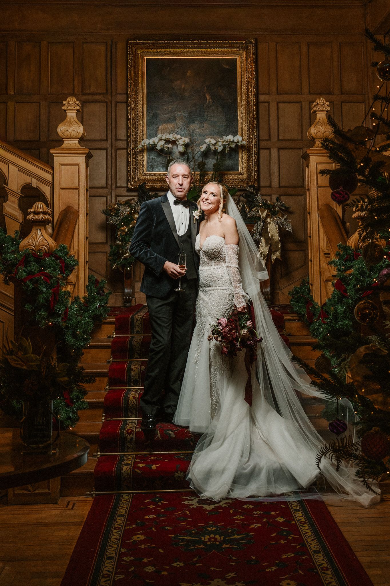 Wedding couple poses on a classic wooden staircase with red carpet and holiday decorations.