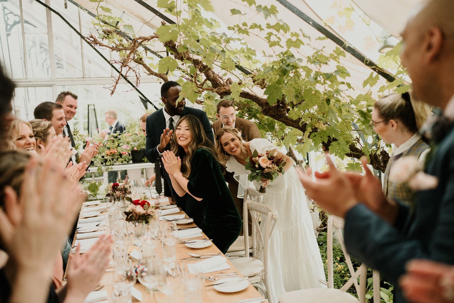 Wedding guests celebrate at an elegant greenhouse reception with hanging greenery and table settings.