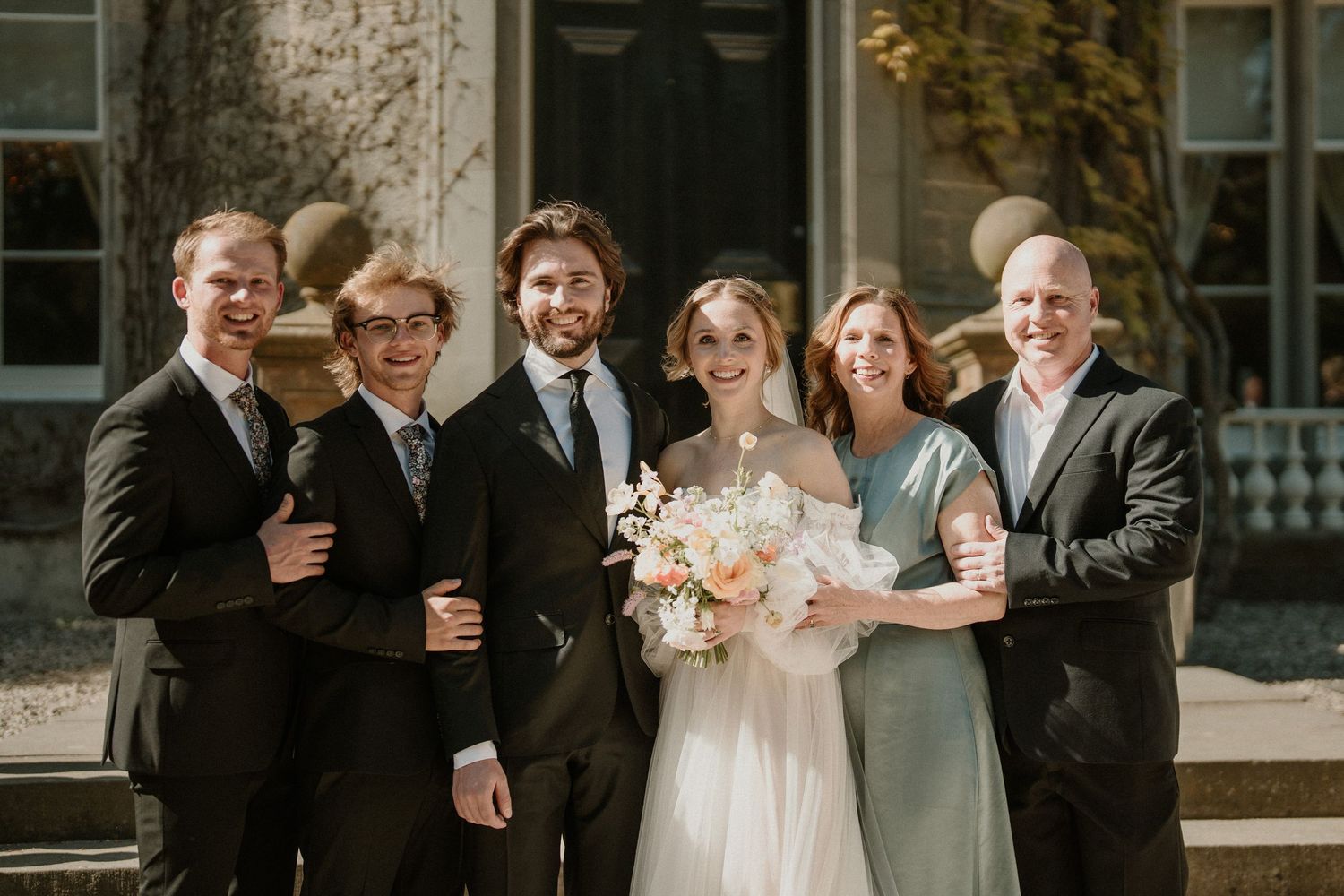 Wedding party gathering outside a stone mansion on a sunny day.
