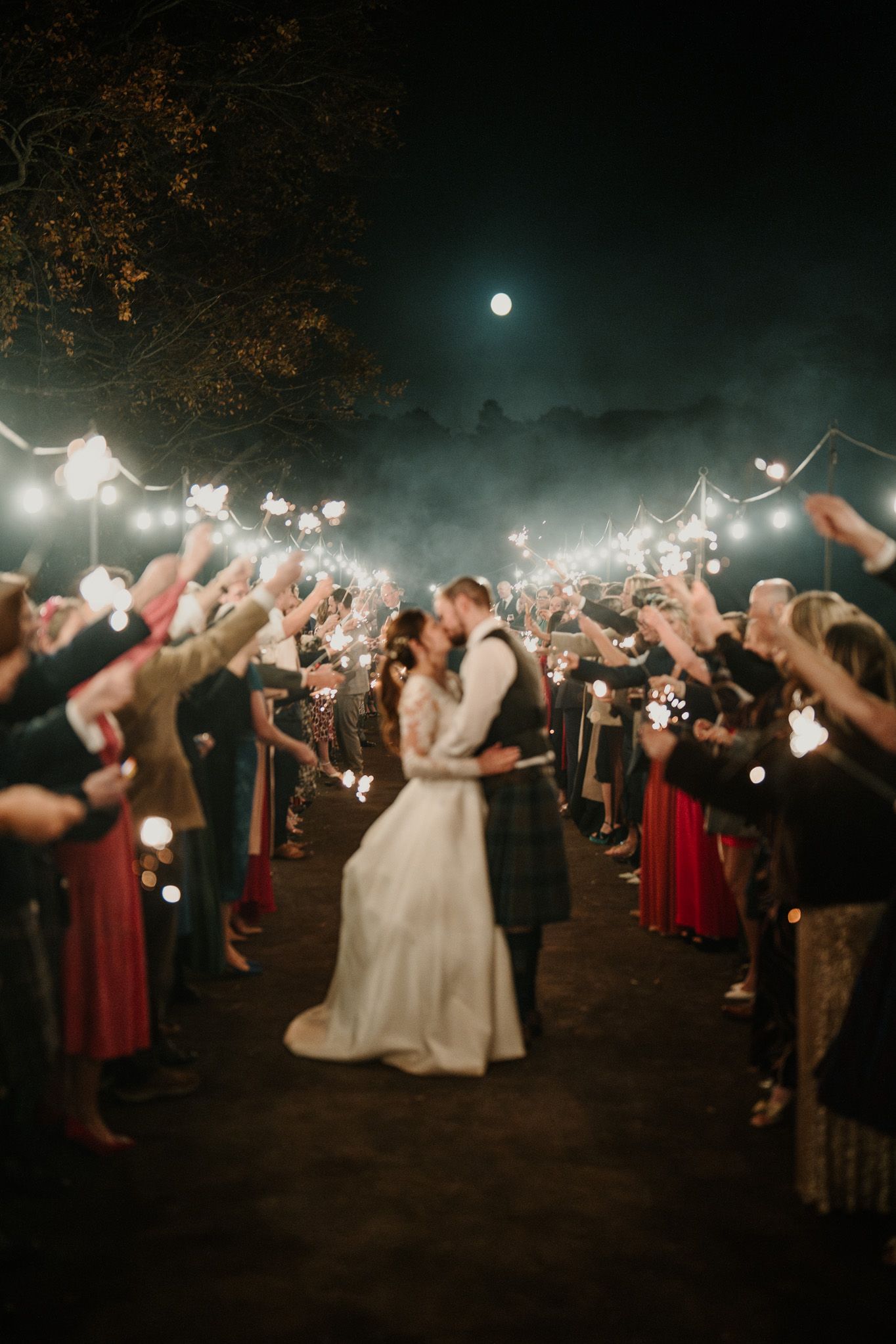 Wedding party and guests form a sparkler tunnel under the night sky with string lights overhead.