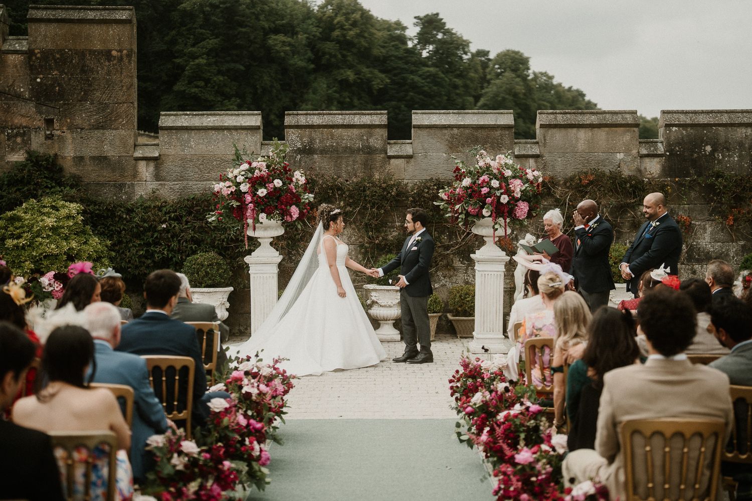 Elegant outdoor wedding ceremony setup with stone walls and floral arrangements in pink and burgundy tones.