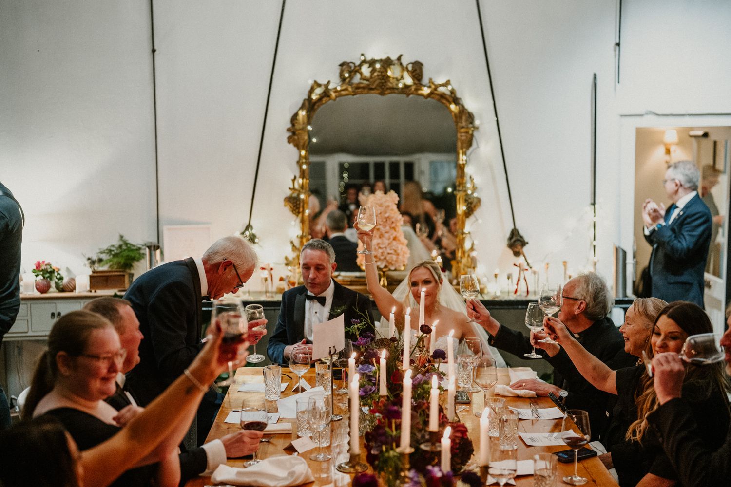 Wedding guests enjoy an elegant candlelit dinner reception with ornate gold mirrors and warm lighting.