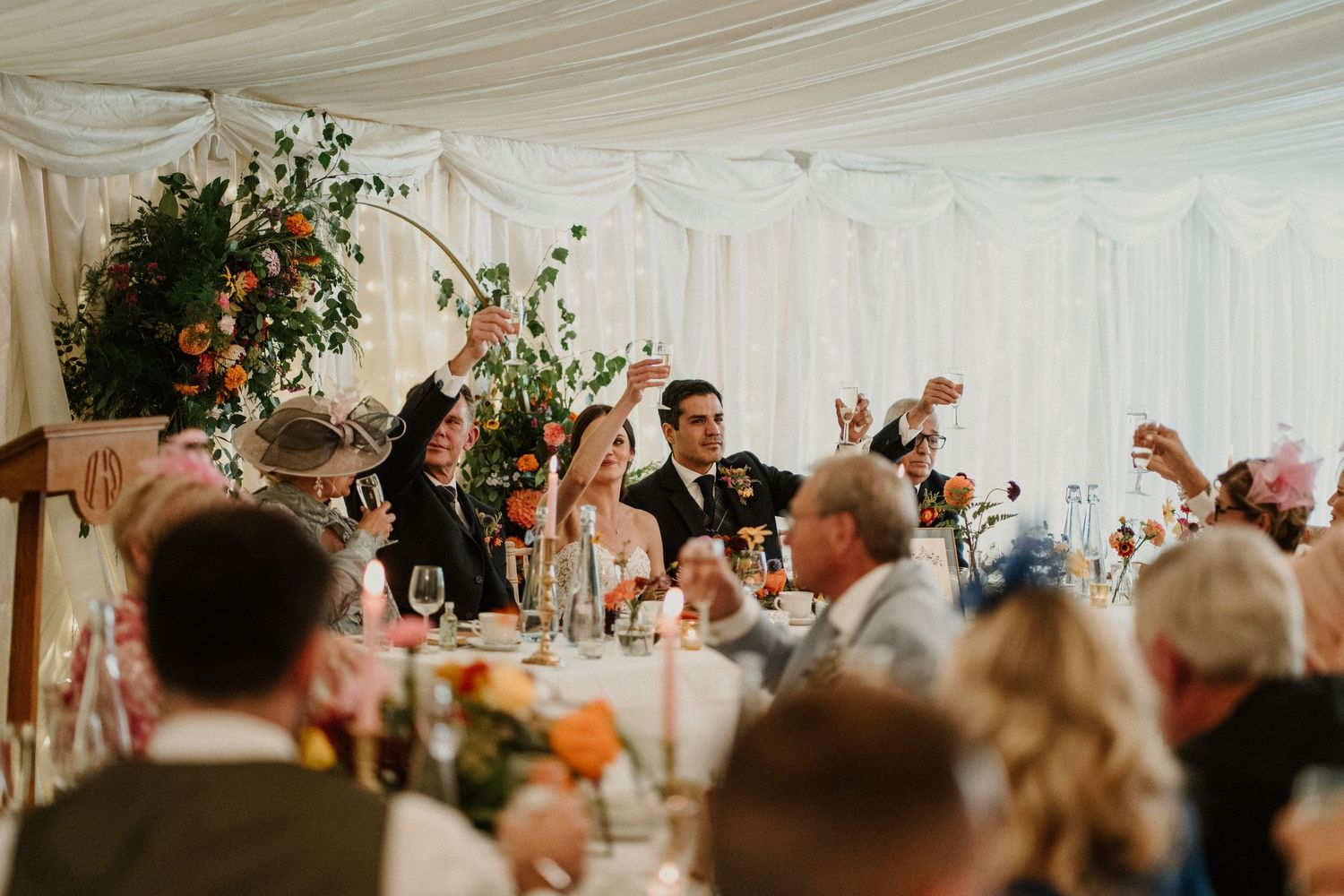 Wedding guests raise glasses for a toast inside an elegantly decorated white marquee tent.