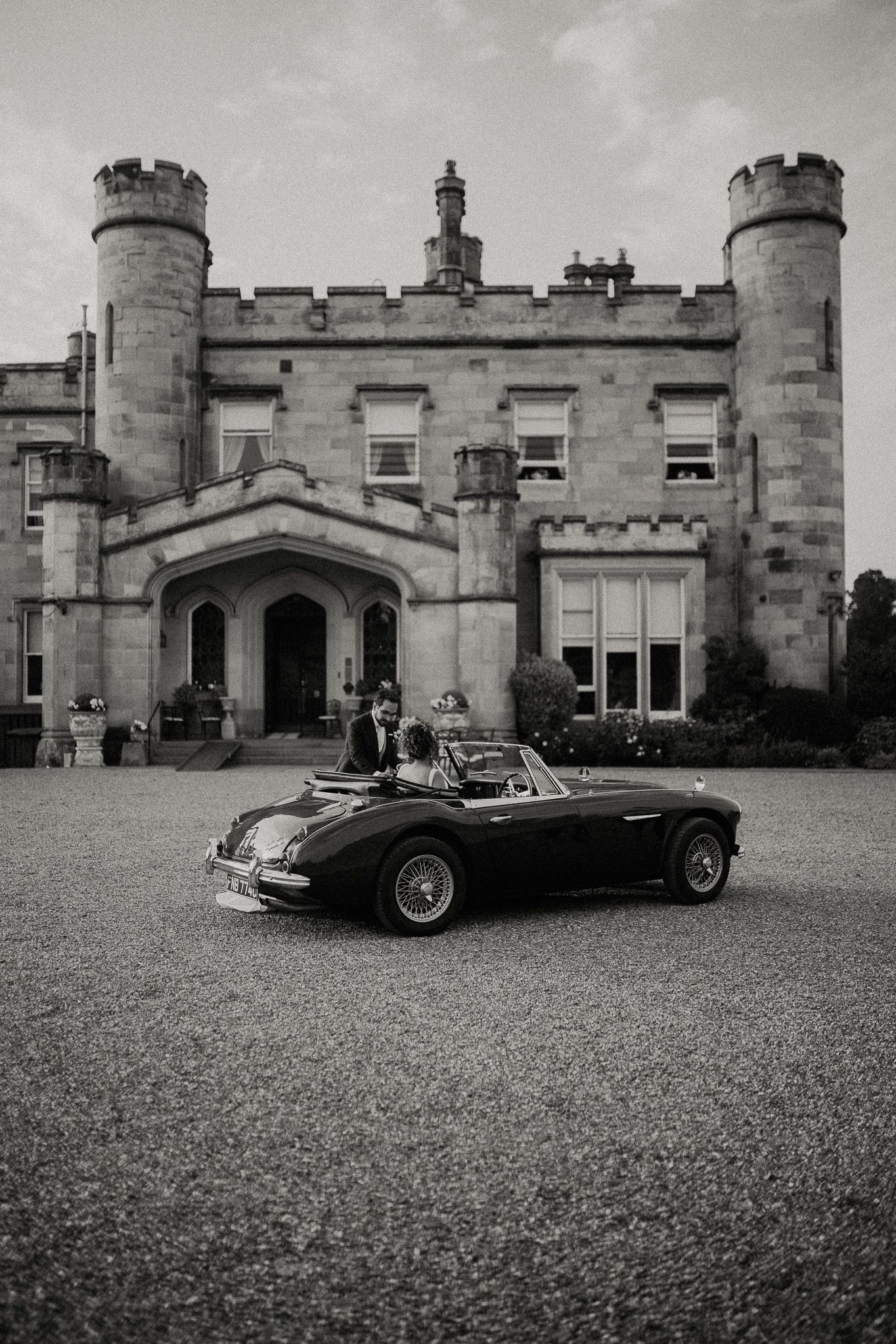 Black and white photograph of a vintage sports car parked in front of a Gothic castle estate.