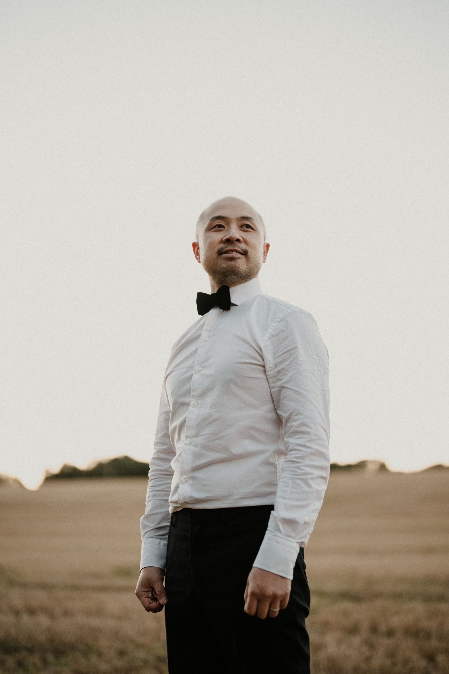 A person in formal attire poses against a sunset sky in a field setting.