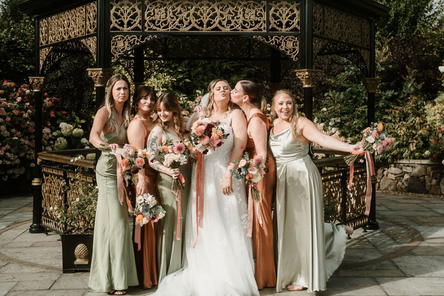 Bridesmaids in sage and rust dresses posing with bride under garden pavilion.