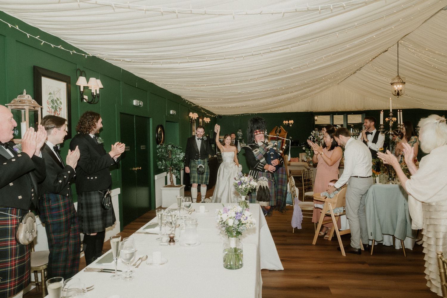 Bride and groom enter wedding reception hall to applause, guests in kilts celebrate in elegant green marquee.