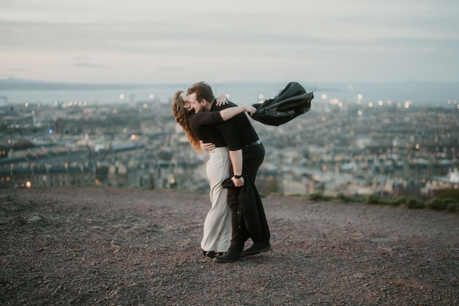 Couple sharing a romantic dip kiss on a hilltop overlooking a city skyline at dusk.