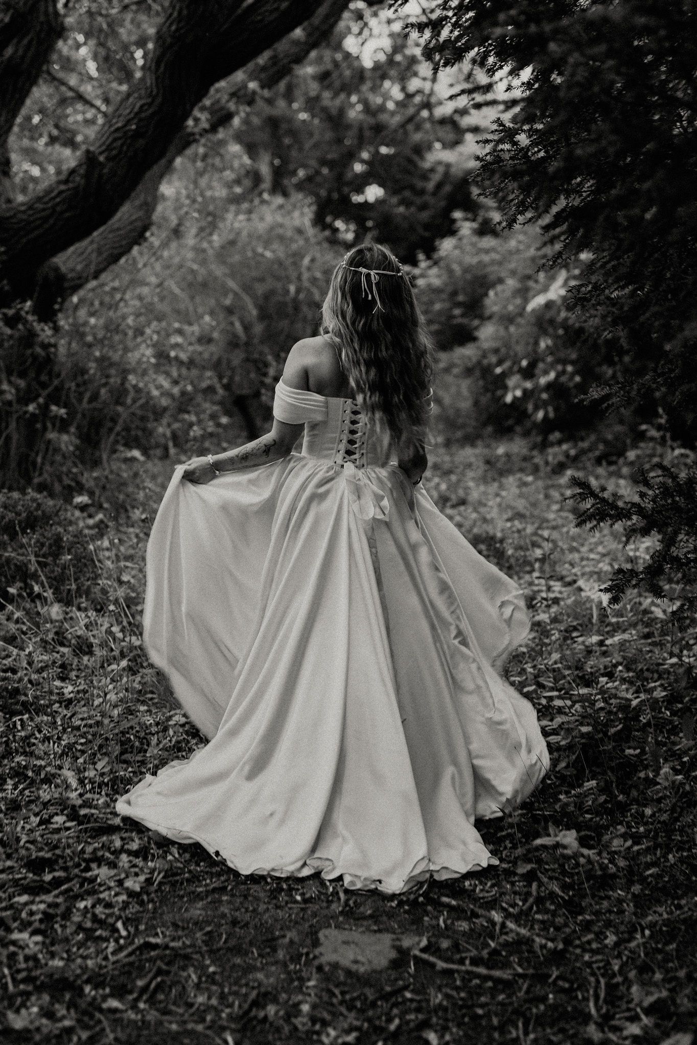 Black and white photo of a bride in an off-shoulder ball gown walking through a lush forest path.