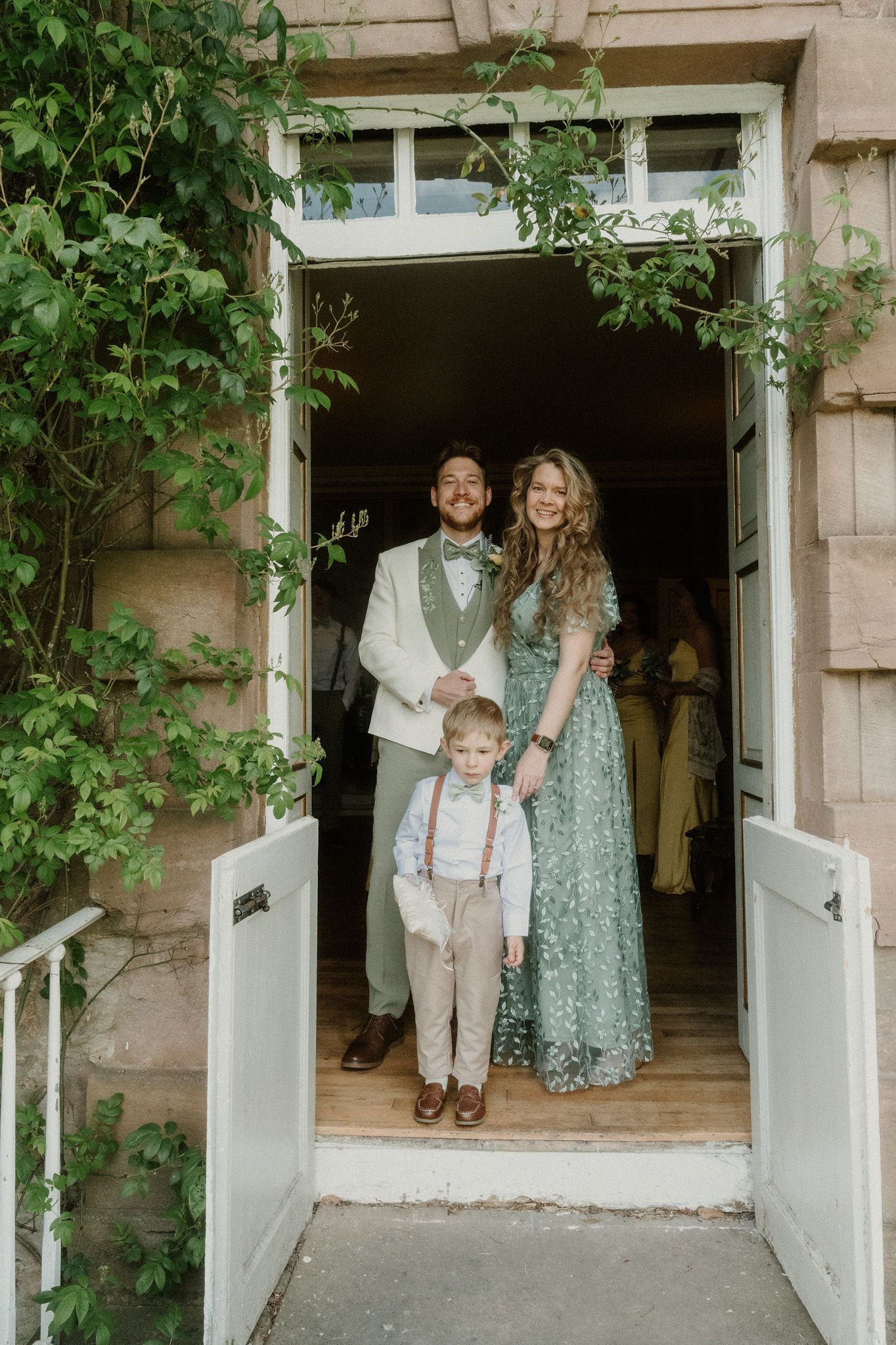 Bride in green floral dress and groom in cream suit stand in doorway with young boy in suspenders.