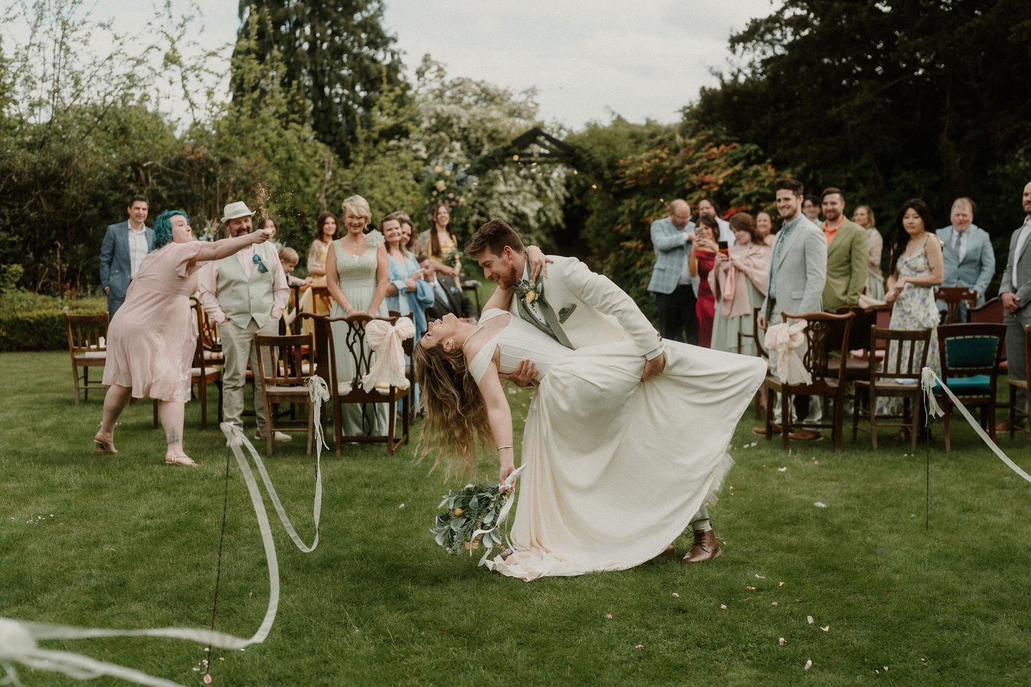 Bride and groom share romantic dip kiss during outdoor wedding ceremony on lush green lawn with guests celebrating.