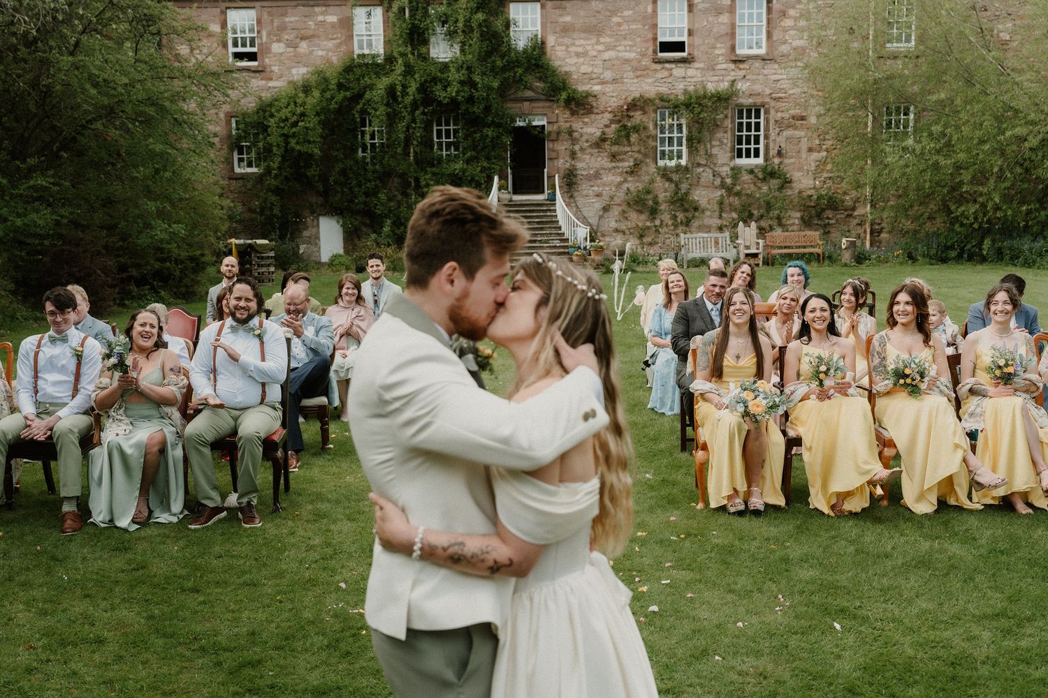 Newlyweds share first kiss at outdoor wedding ceremony with bridesmaids in yellow dresses and guests seated on lawn.