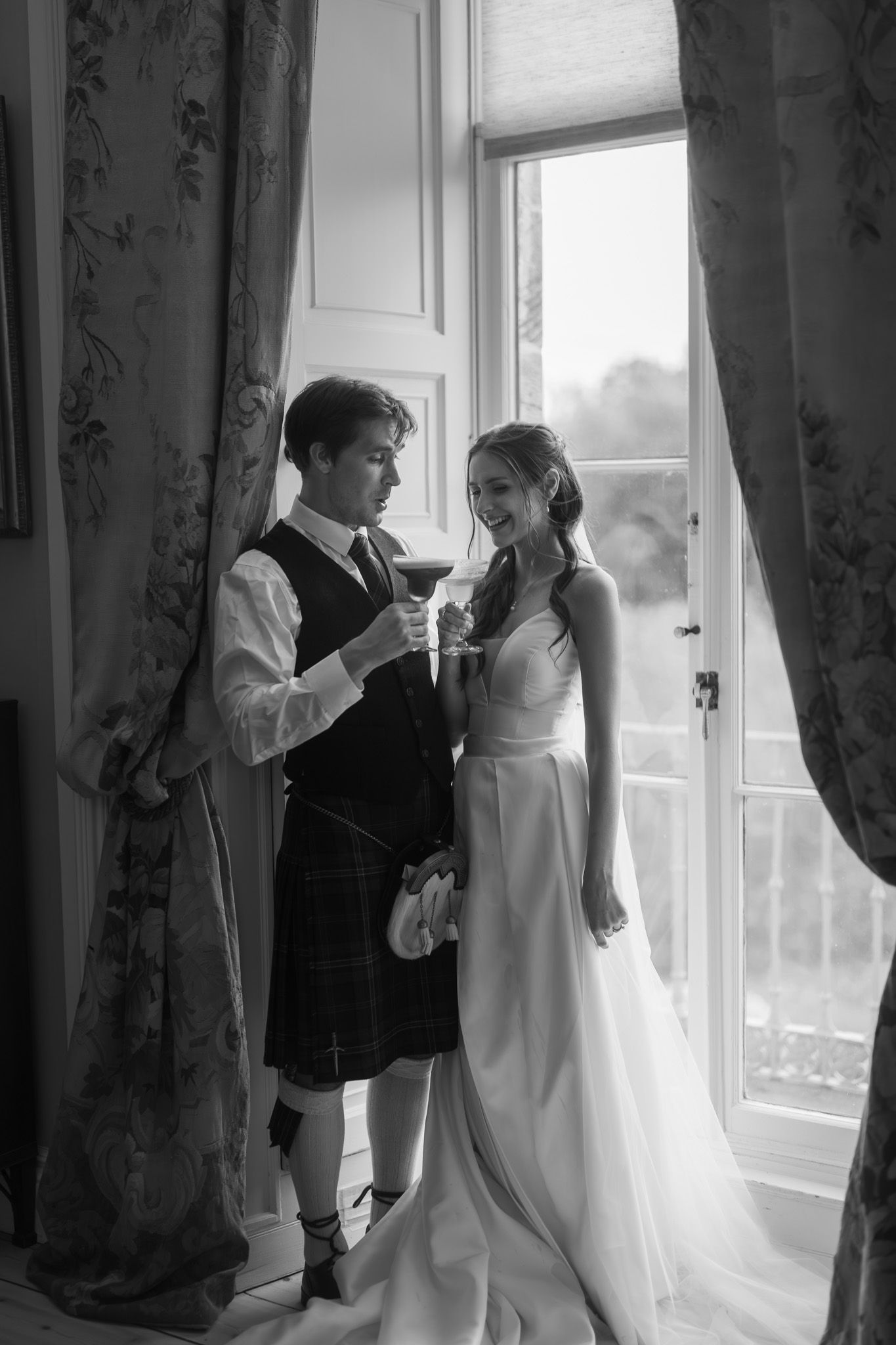 Black and white photo of a groom in a kilt and bride in a white gown sharing a toast by a sunlit window.