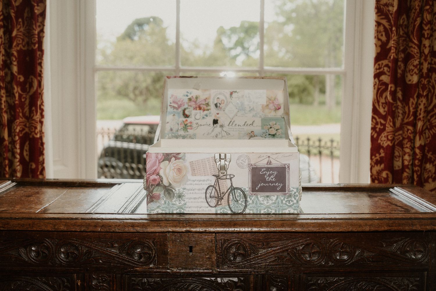 Decorative floral suitcase wedding card box displayed on antique wooden dresser near window.